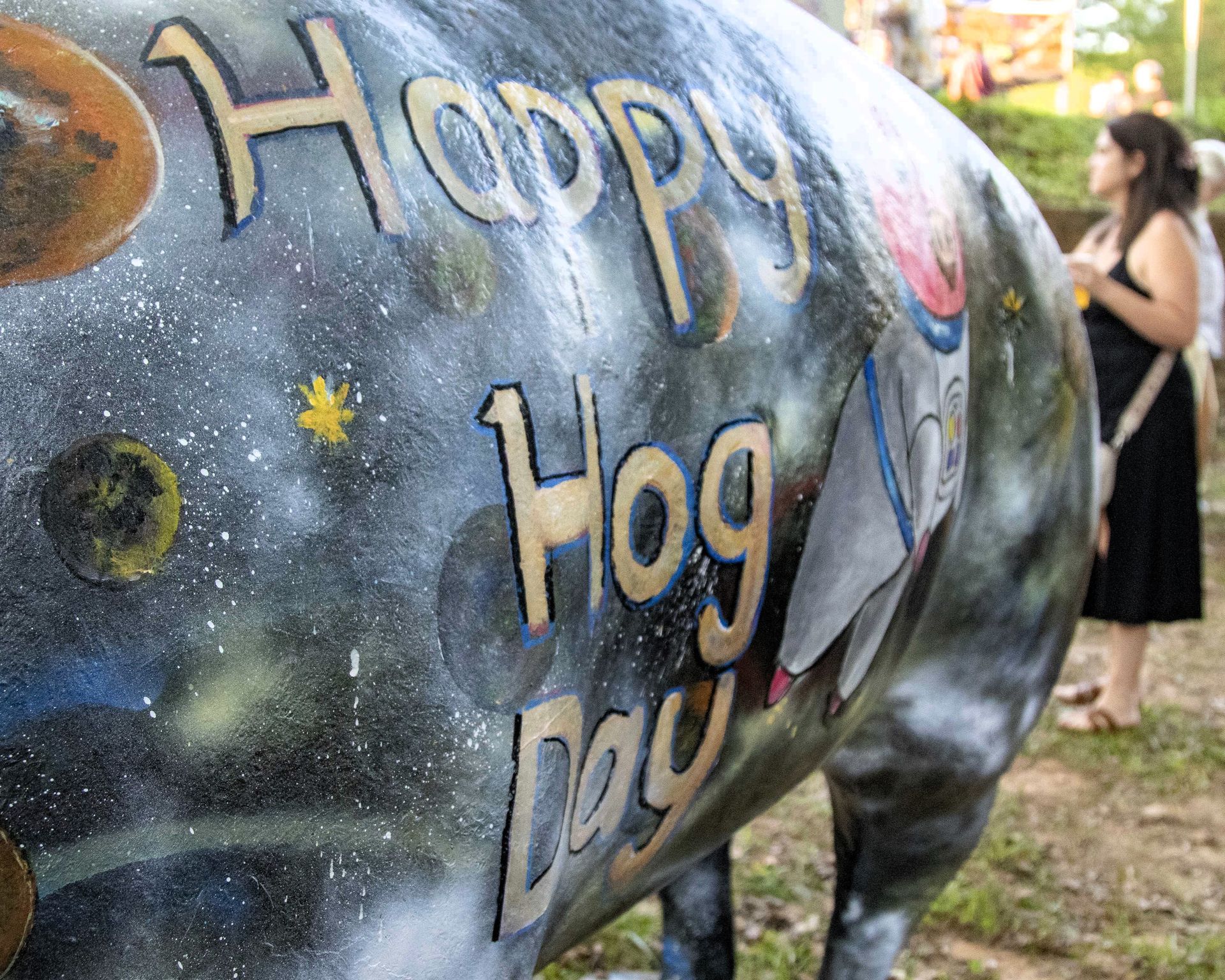 Painted pig sculpture with “Happy Hog Day” text outdoors; woman stands in the background.