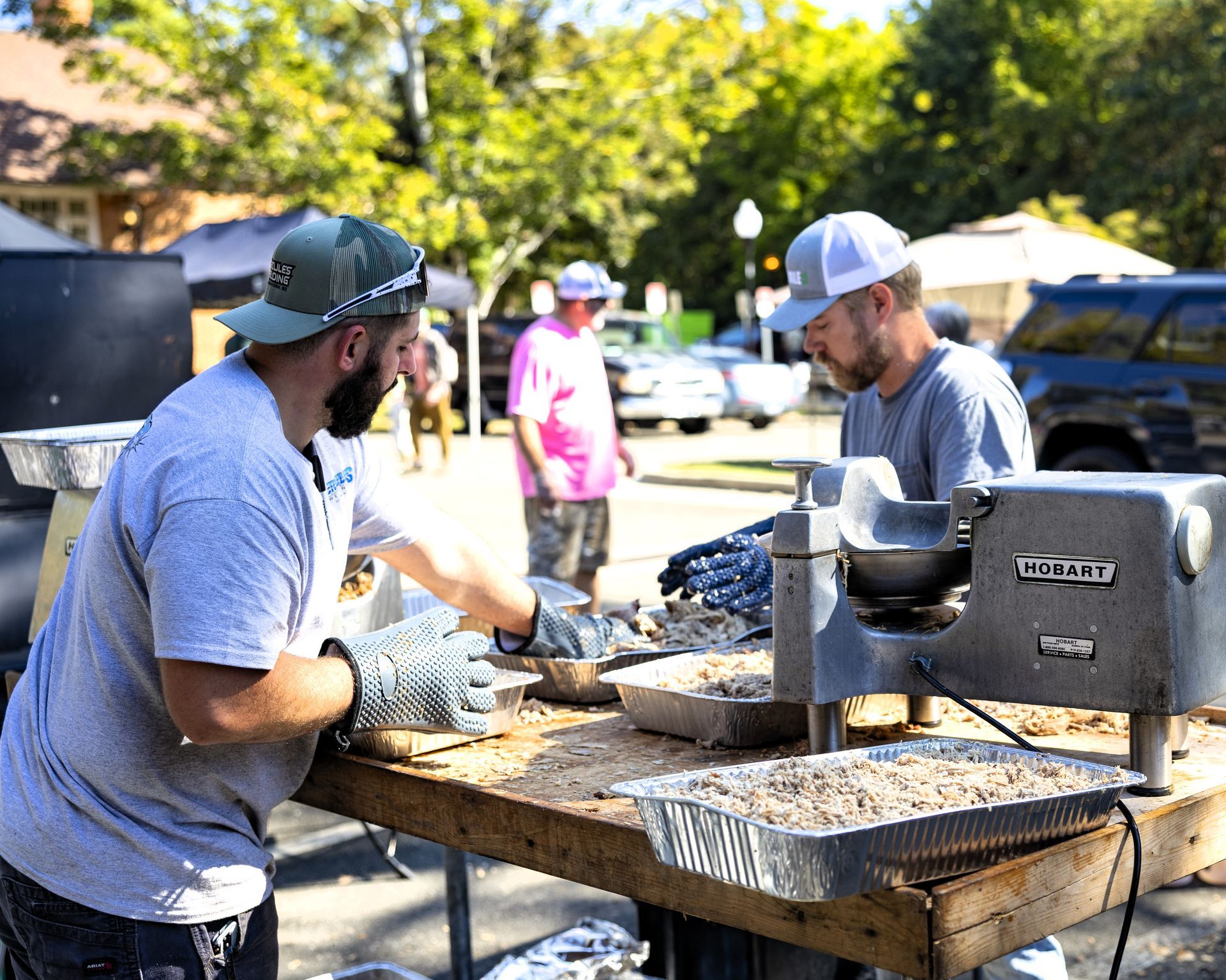 Two workers in caps and gloves shaping material at a table outdoors on a sunny day