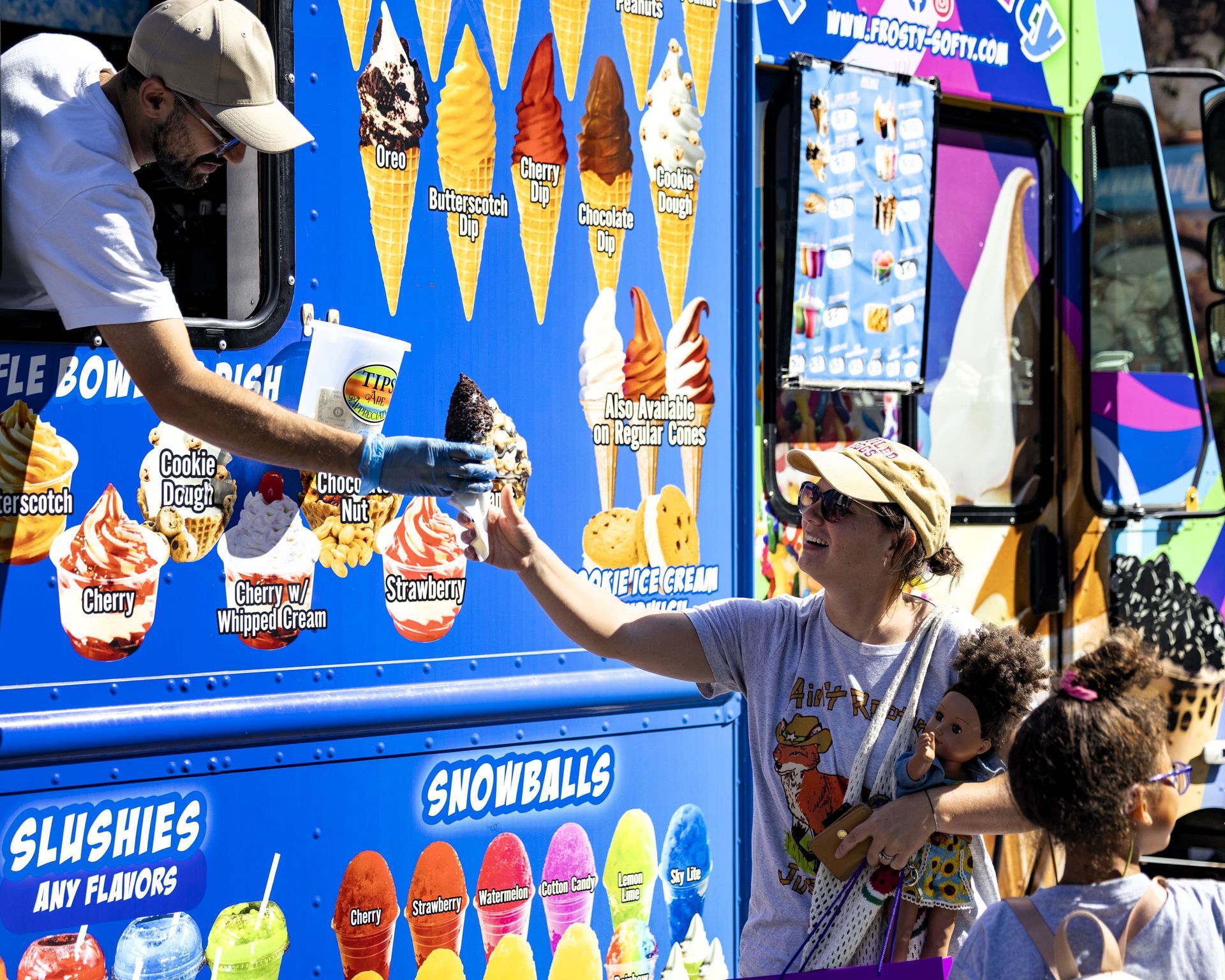 Vendor handing a blue cup from an ice cream truck to a customer at a sunny street fair