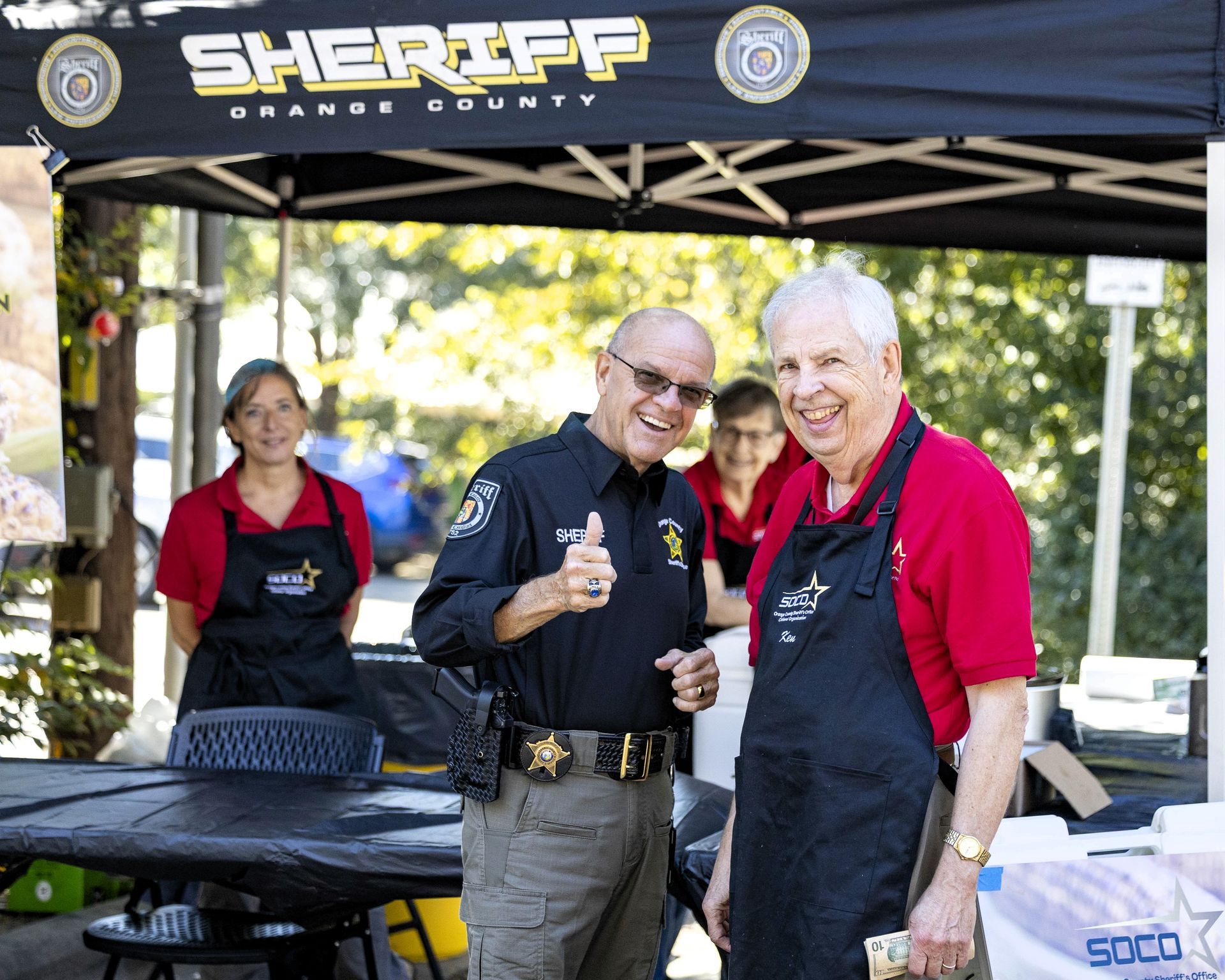 Sheriff’s booth with three volunteers standing under a canopy, one holding a cup and smiling