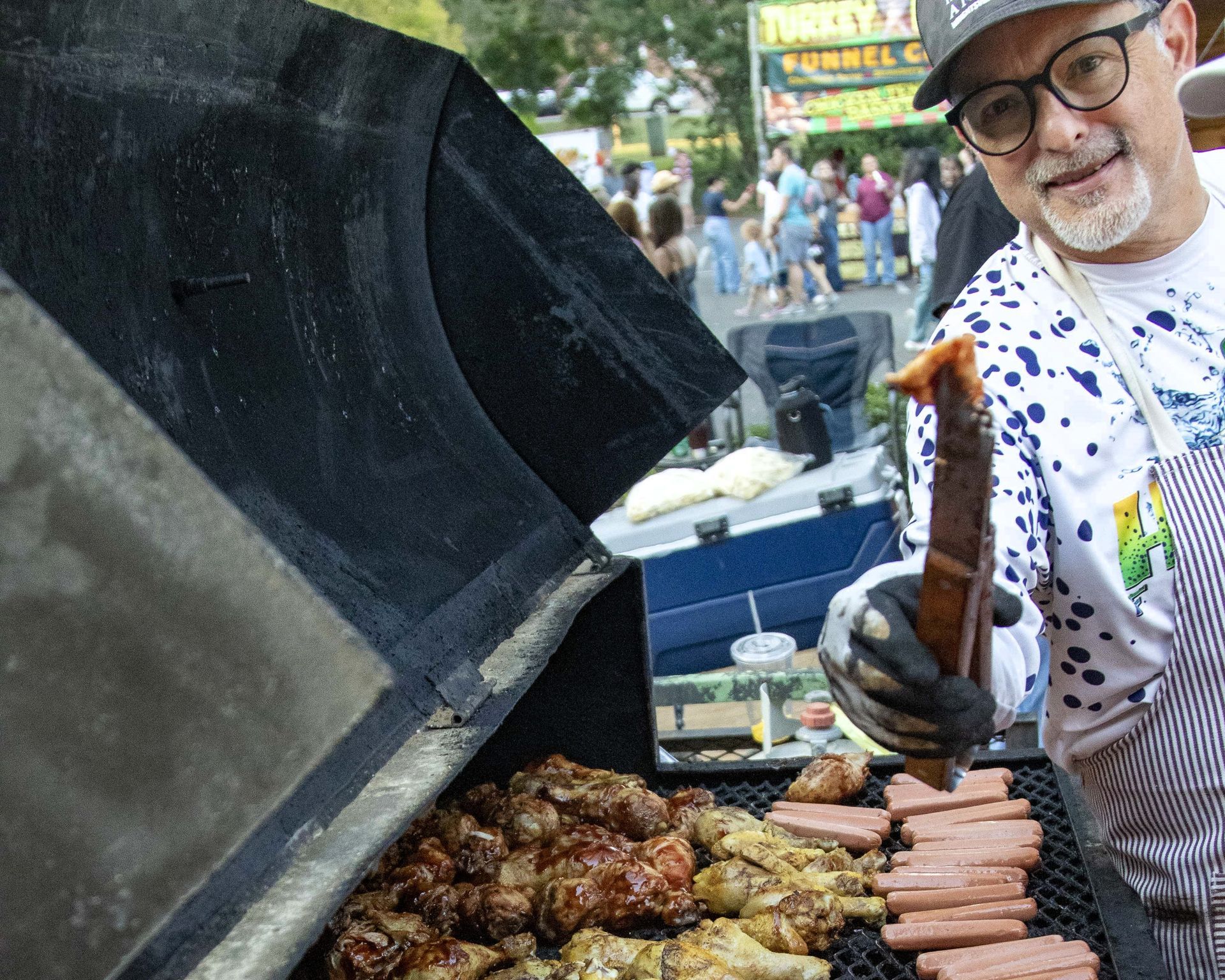 Man grilling sausages and meat at an outdoor market, smiling beside a large barbecue grill.