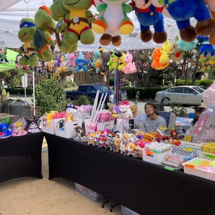 Outdoor market stall with hanging plush toys and colorful trinkets; vendor seated behind tables displaying toys.