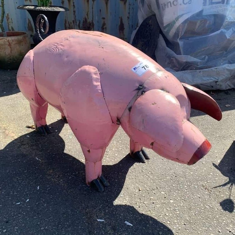 Pink pig sculpture made of metal or wood, standing outdoors on pavement in sunlight
