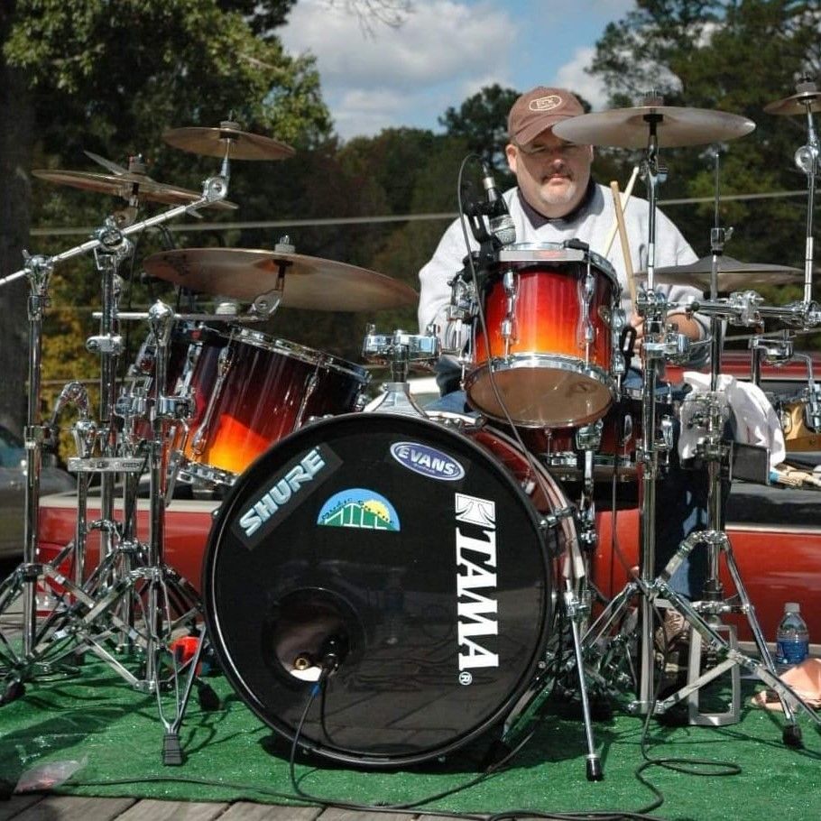 Drummer seated behind a red-orange drum kit outdoors on a green mat, wearing a cap and sunglasses.