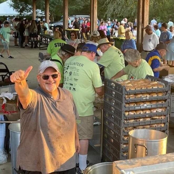 Man smiling and pointing at a crowded outdoor food-serving area with trays and volunteers.