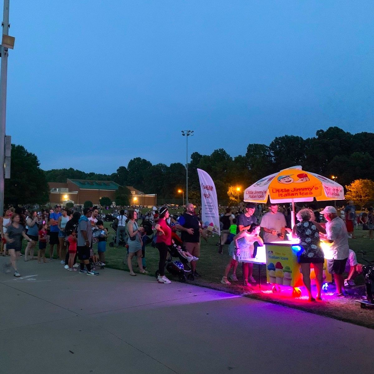 Crowd gathered at dusk beside a glowing neon-lit vendor booth at an outdoor event