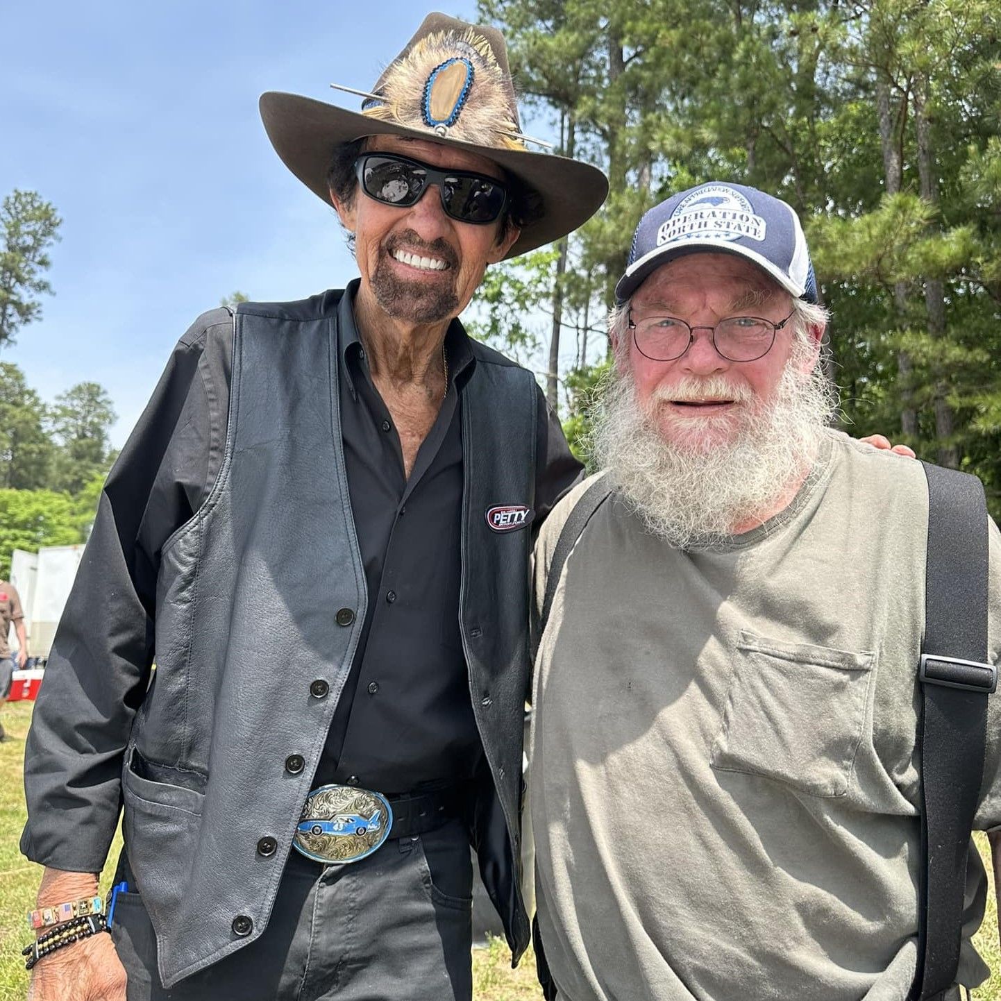 Two smiling men posing outdoors, one in a black hat and one in a blue cap, with trees in the background