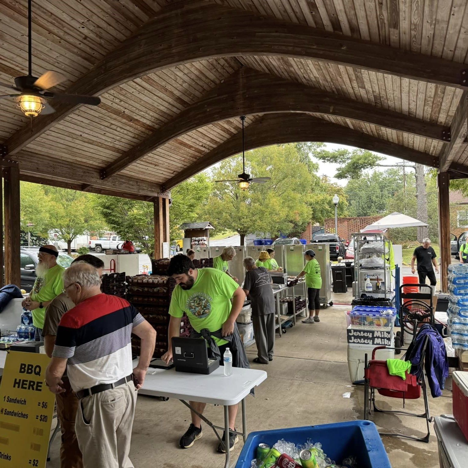 Open-air BBQ gathering under a pavilion with people standing near grills and food tables.