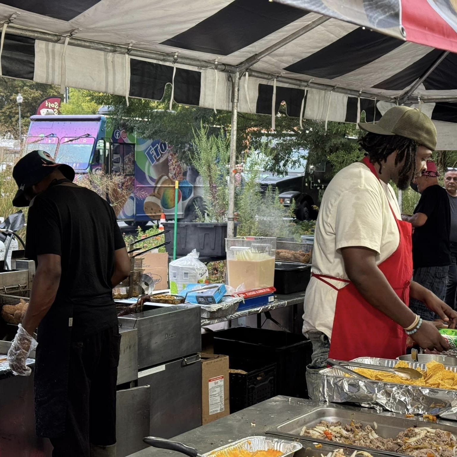 People serving food at an outdoor stall under a canopy, with trays and cooking equipment.