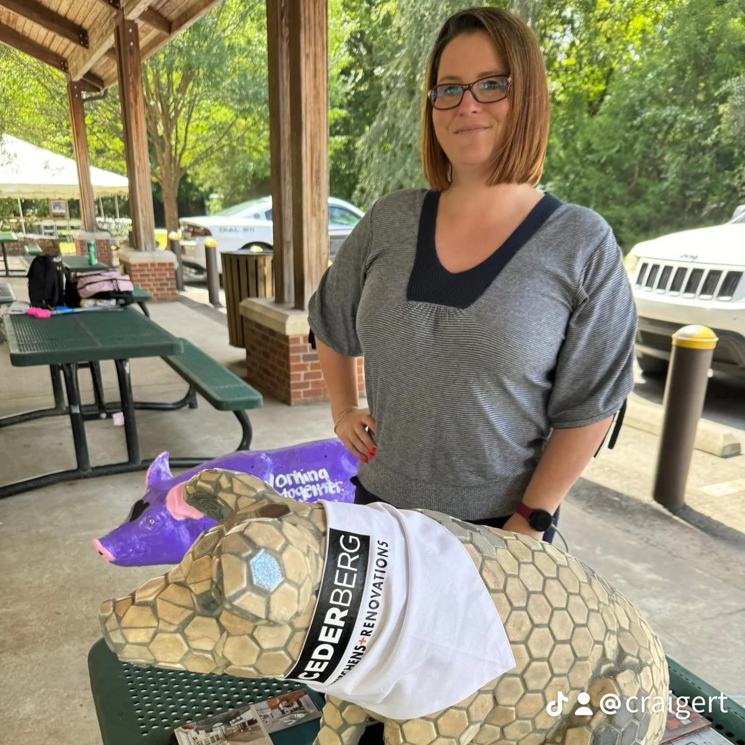 Woman standing beside a large tortoise at a picnic shelter, wearing a gray top and glasses.