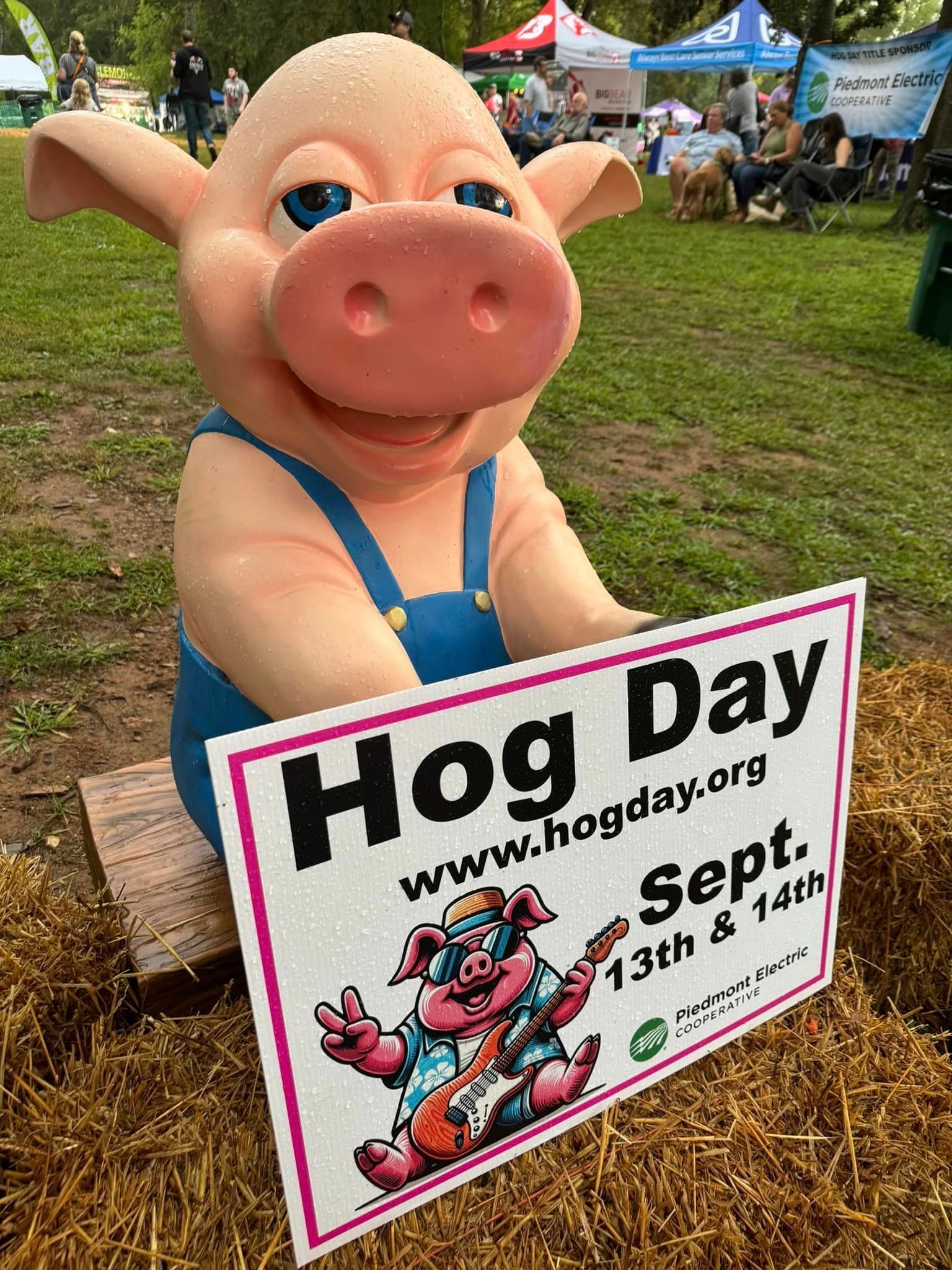Pink pig statue with “Hog Day” sign at an outdoor fair, seated on grass.