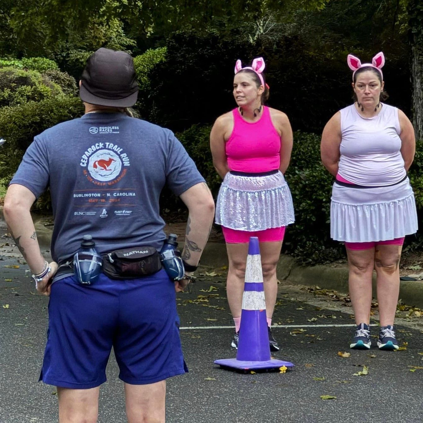 Two women in pink and gray outfits with cat ears stand outdoors facing a person in blue shorts and hat.