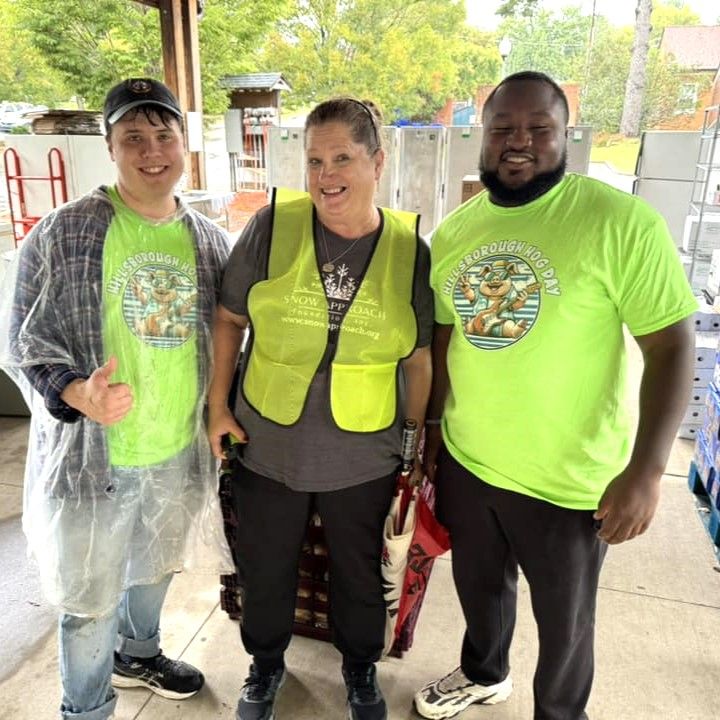 Three people in neon shirts and safety vests stand smiling under a pavilion.