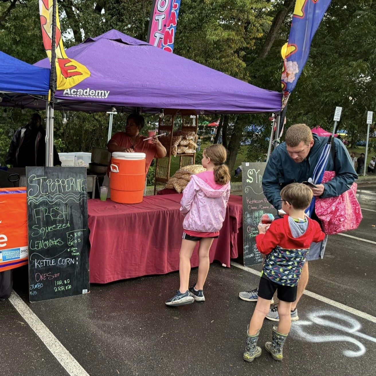 People at a purple-tented outdoor market stall on a rainy day, with two children waiting at the counter.