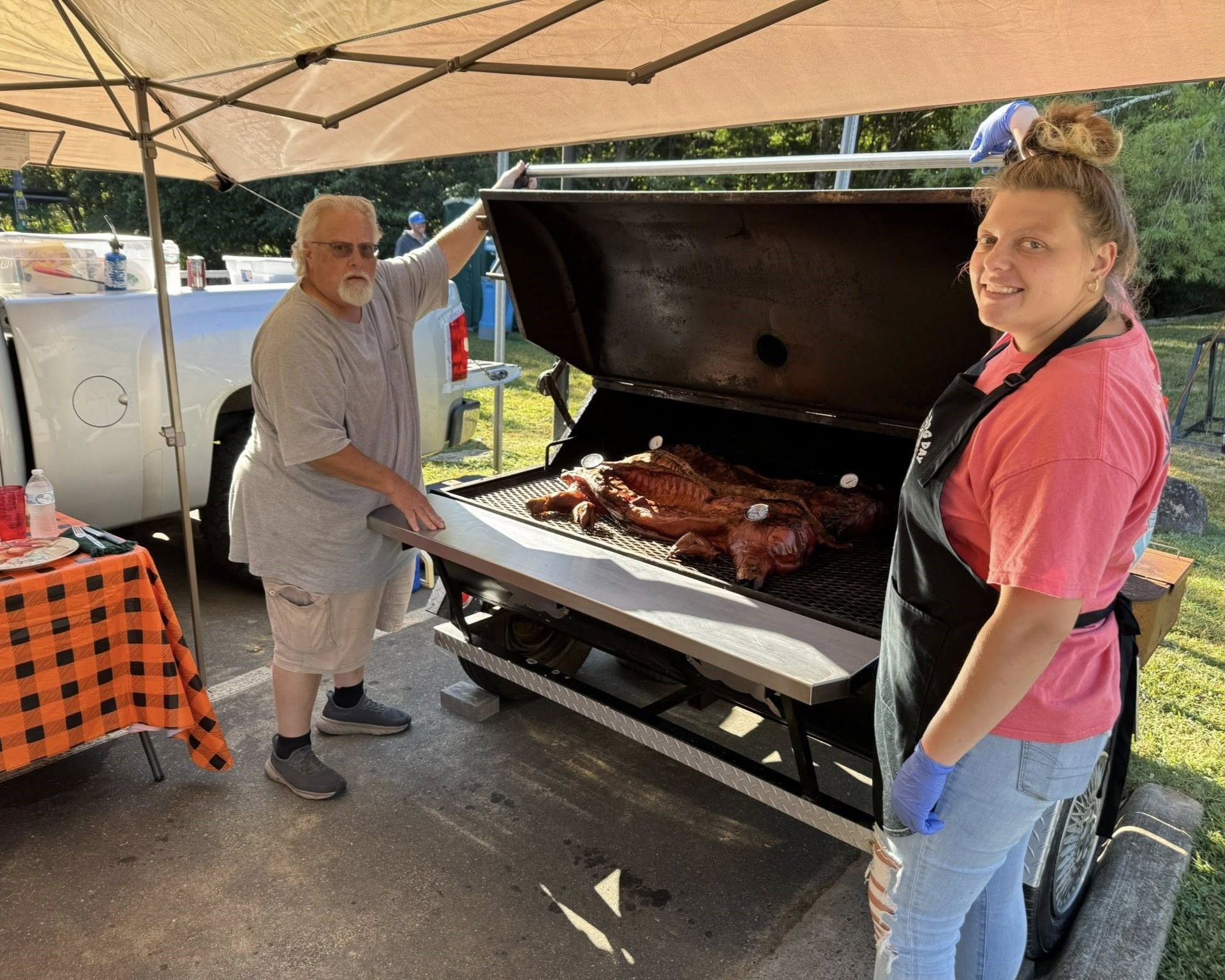 Two people stand by a large open barbecue grill with smoked meat under a tent outdoors.