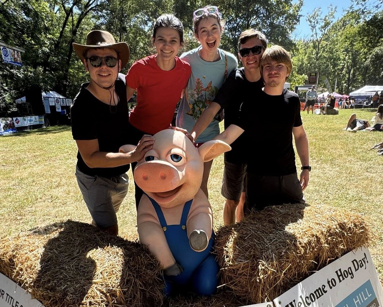 Group posing with a pig mascot in a sunny outdoor park, with hay bales and trees in the background