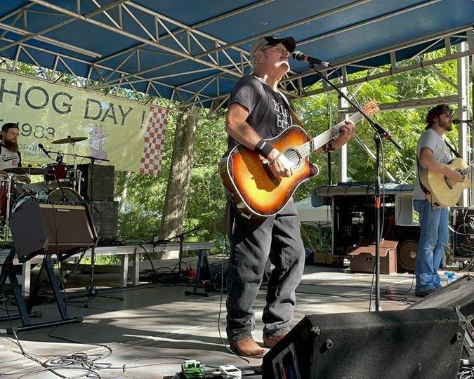 Outdoor stage with two musicians playing guitars under a blue canopy, one singing into a microphone.