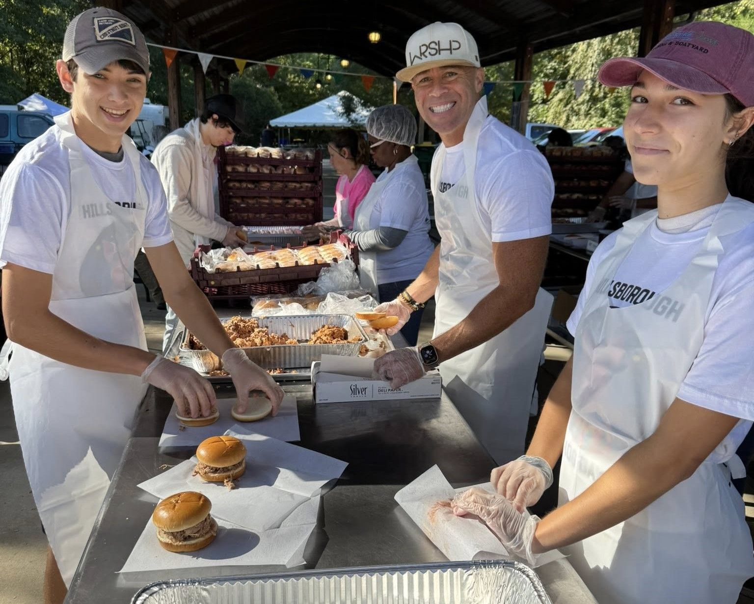 Three volunteers in white shirts serve food at an outdoor buffet table, smiling at the camera.