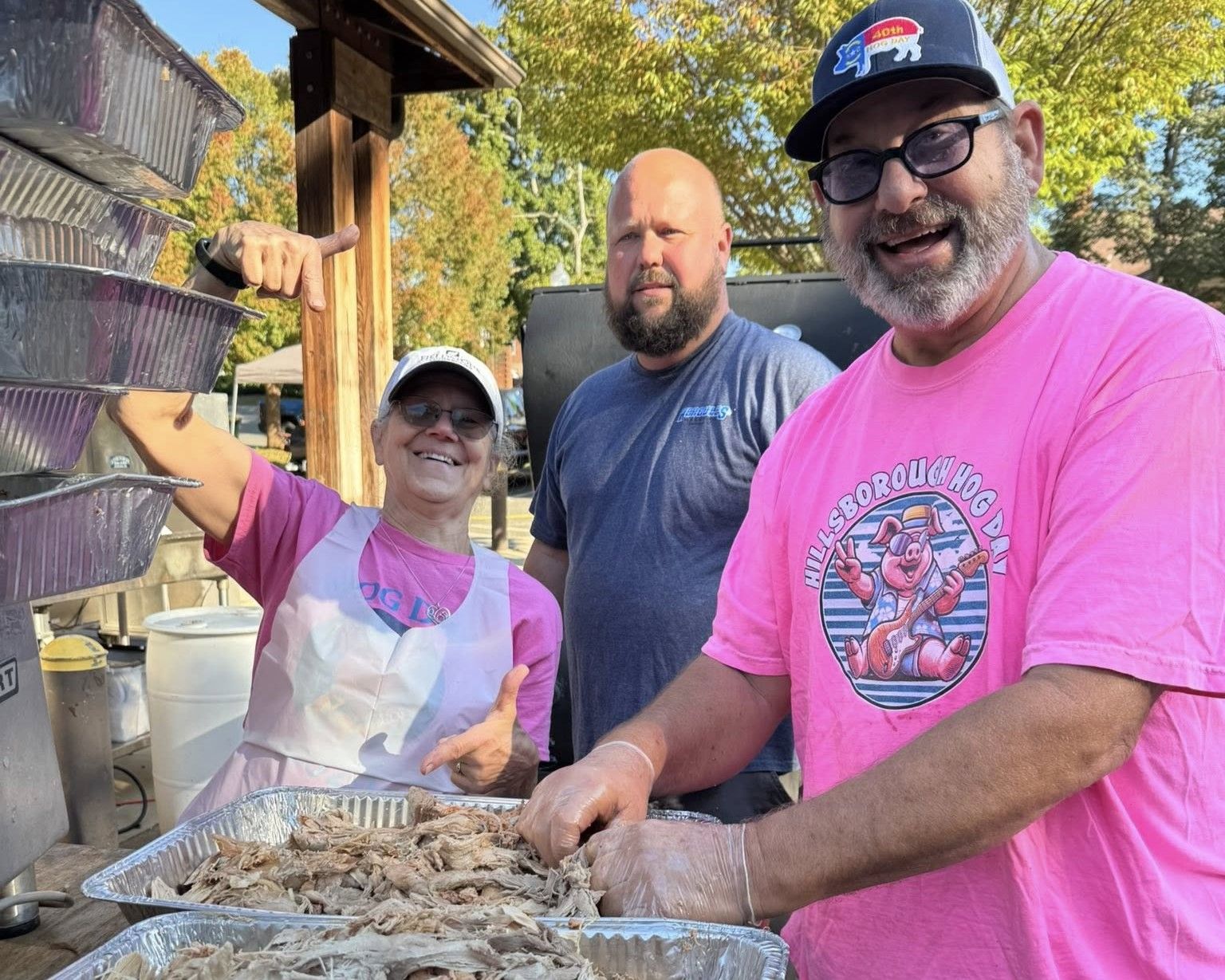 Three people outdoors grilling oysters, smiling beside a table with trays of food.