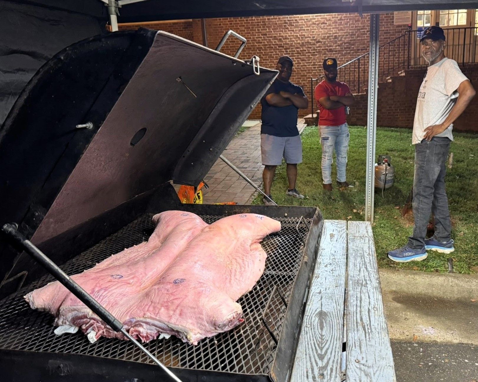 Pig roasting on a grill under a canopy with three people standing nearby at night