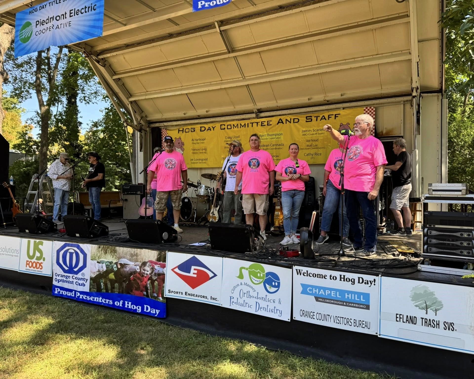 People in bright pink shirts perform on an outdoor stage at a community event with sponsor banners.