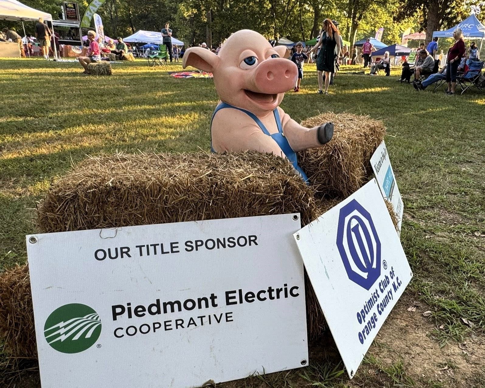 Pig mascot in a cardboard box at a fair, with Piedmont Electric Cooperative sponsor signs.