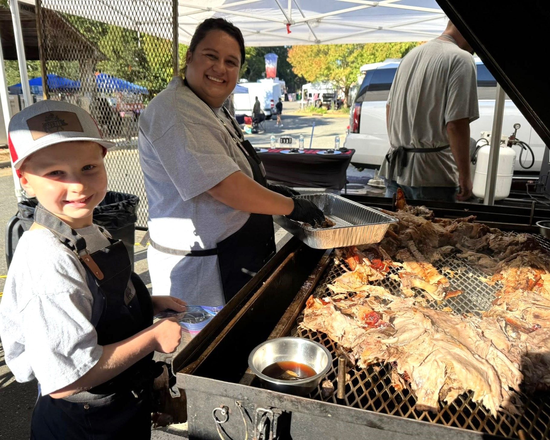 Two people grilling food outdoors at a sunny event, with a large barbecue full of meat and a small sauce bowl