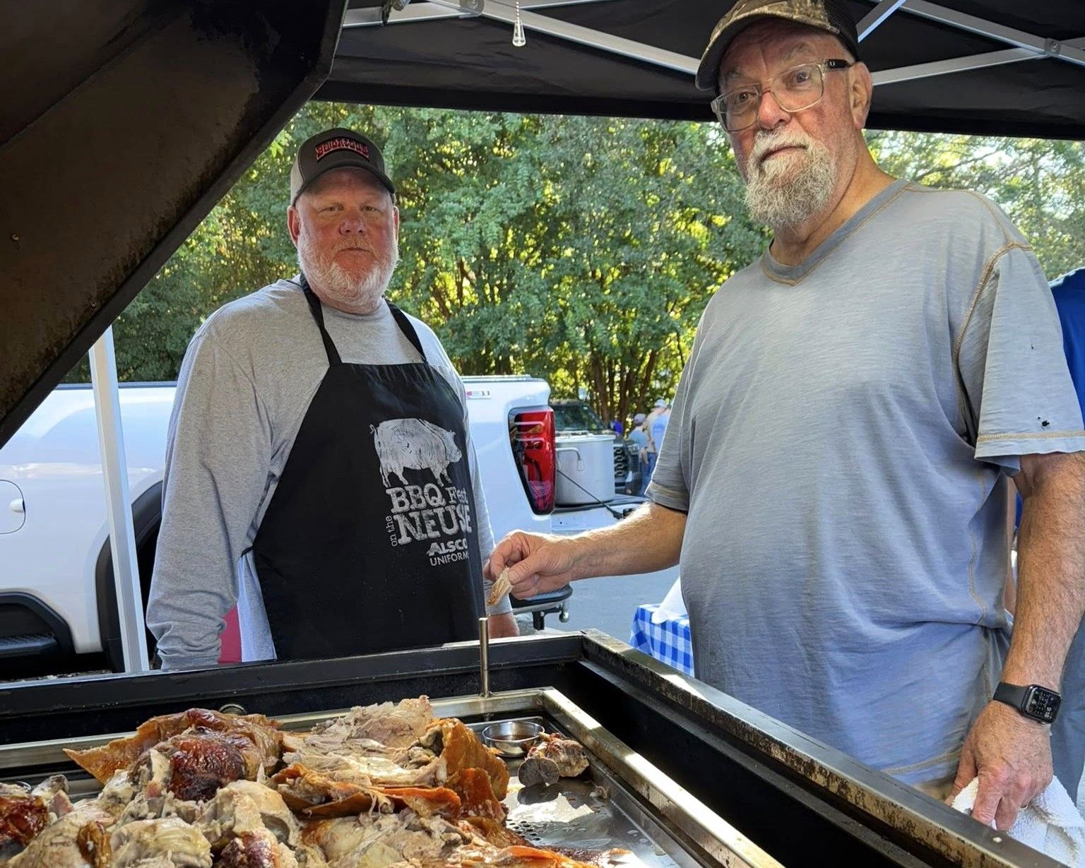 Two men standing by a large outdoor grill with food cooking, smiling under a canopy.