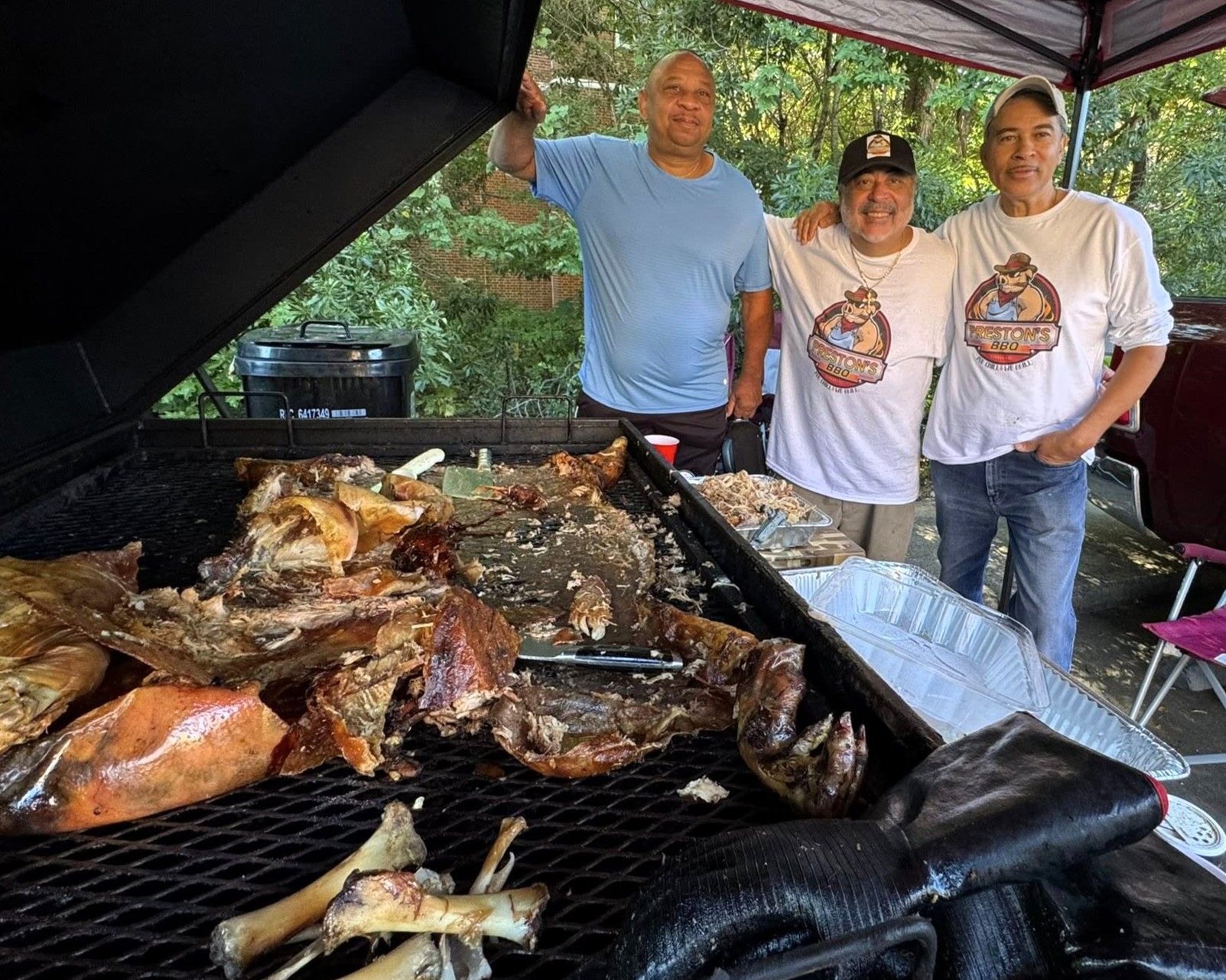 Three people smiling beside a large outdoor barbecue grill piled with assorted meats and fish.