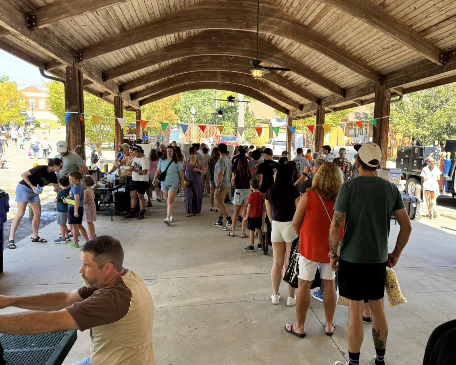 Crowded outdoor pavilion with people mingling and sitting around tables under a wooden roof