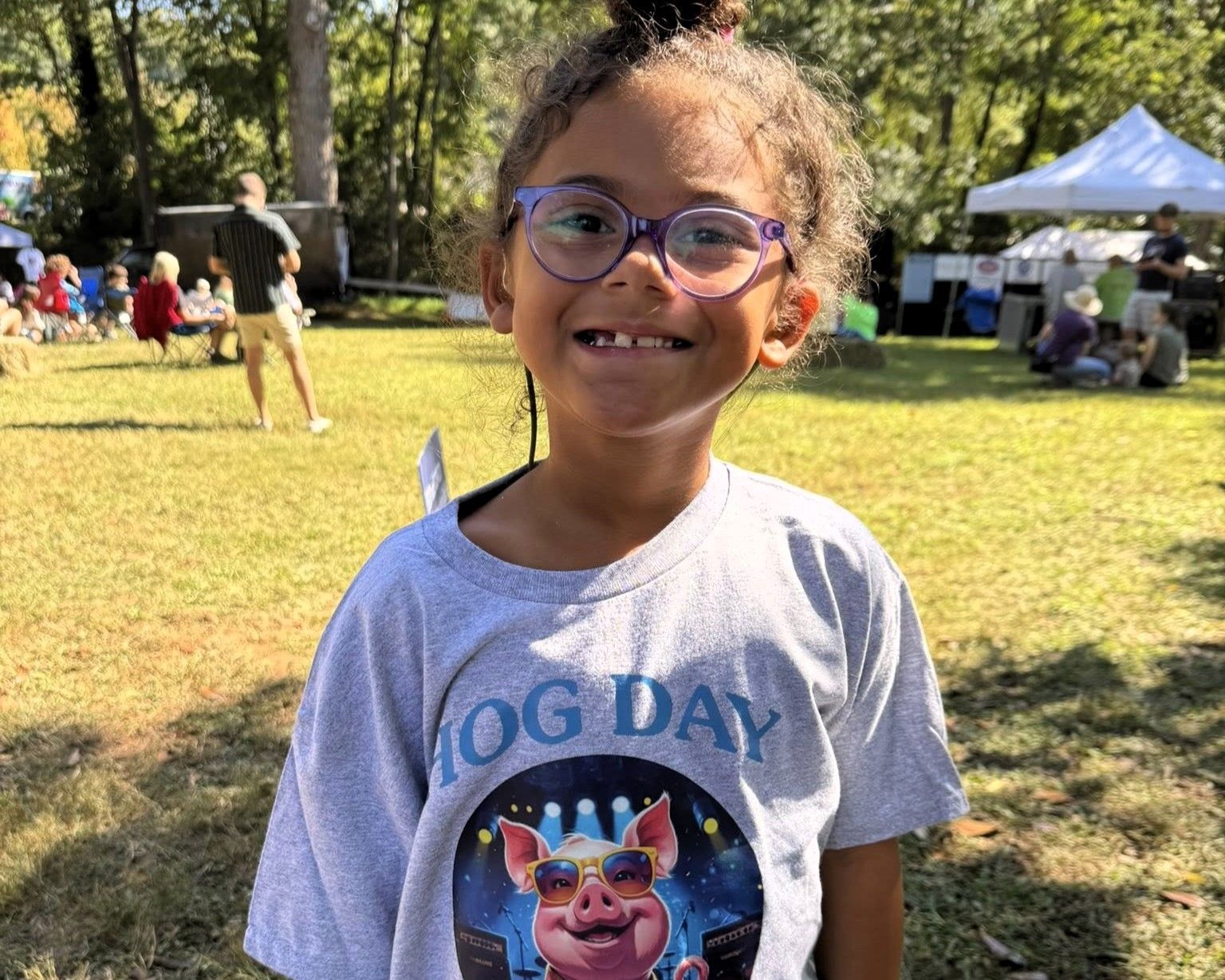 Smiling child in glasses and a “DOG DAY” shirt standing in a sunny park with people and tents behind.
