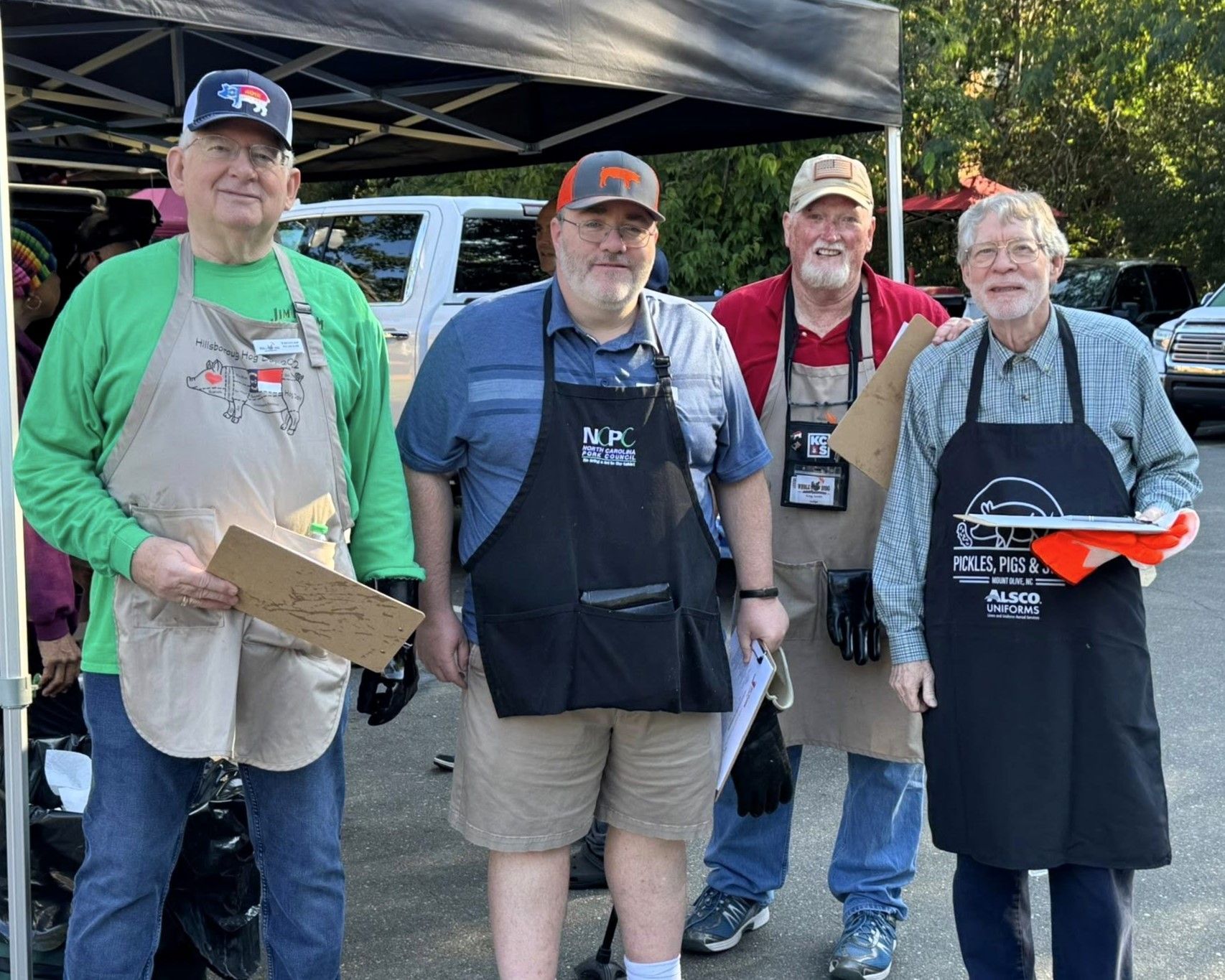 Four people in aprons posing outdoors under a canopy at a food booth.