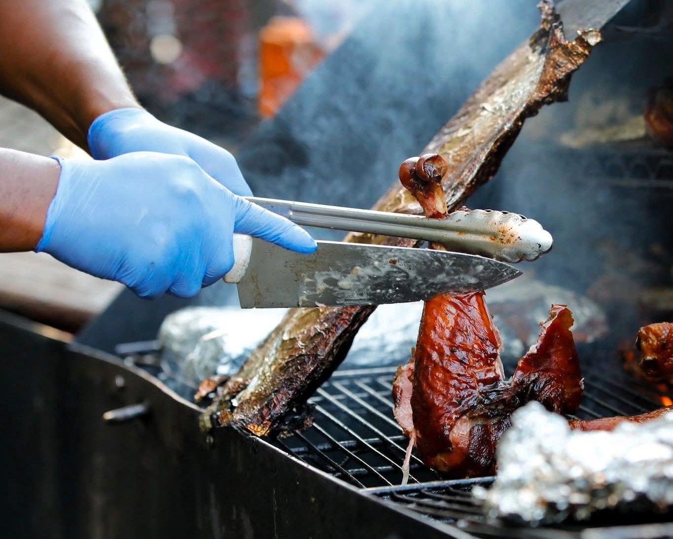 Blue-gloved hands grilling meat with tongs over a smoky outdoor barbecue.