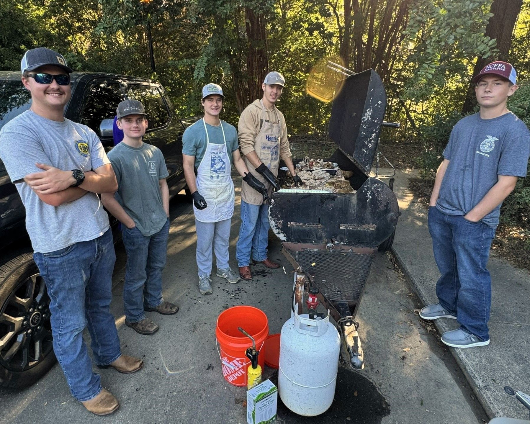Group standing around an outdoor grill with propane tanks and buckets on a driveway, in a wooded area.