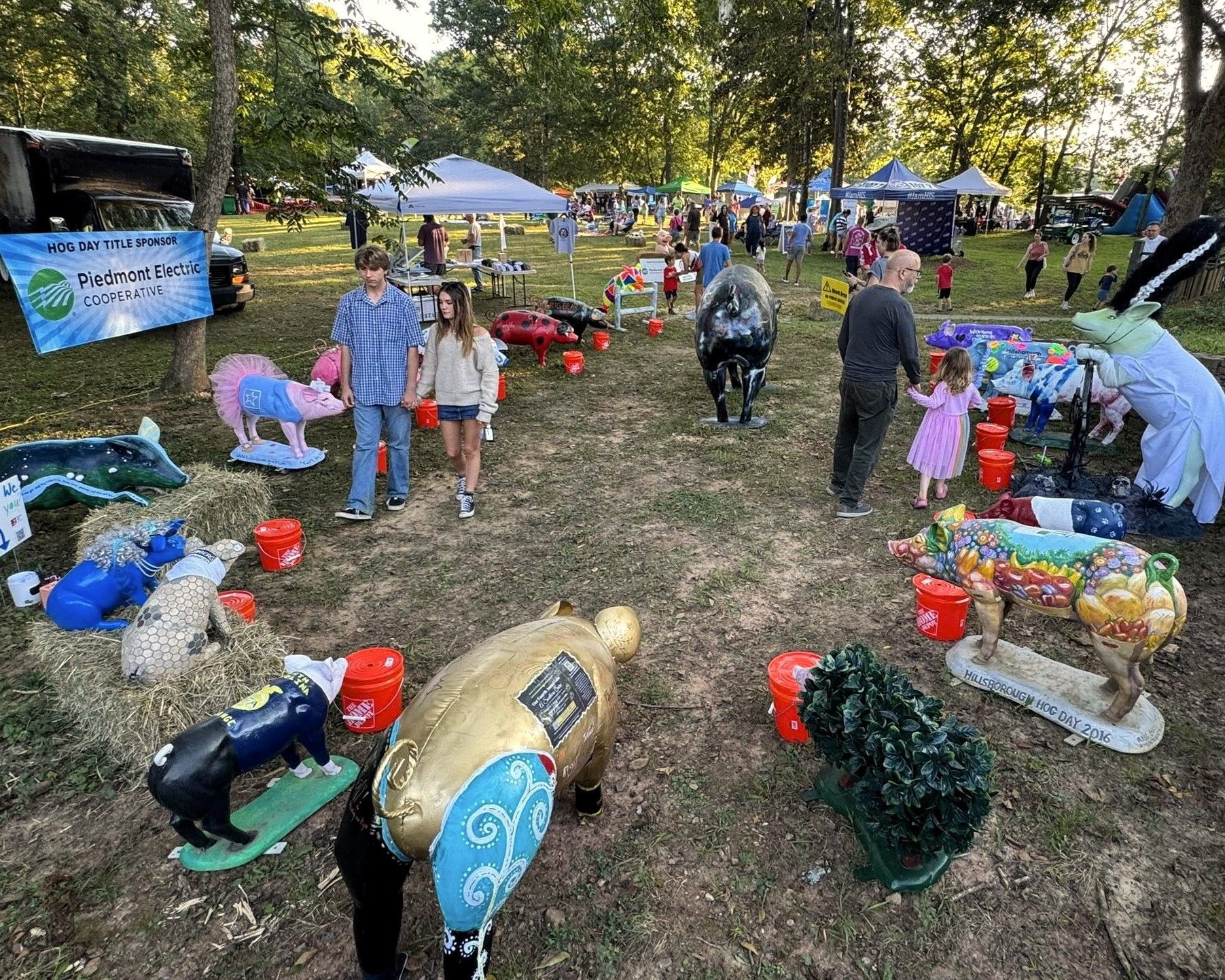 Outdoor art display with colorful painted animal sculptures on grass at a park festival with tents and people