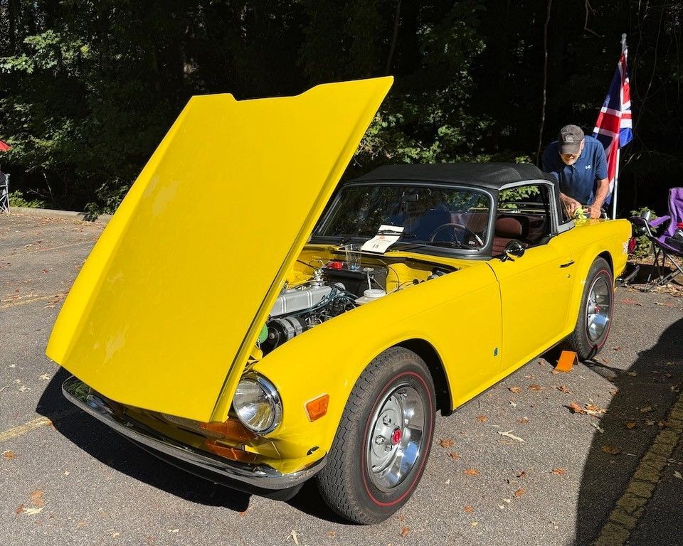 Bright yellow classic convertible with hood open, parked outdoors beside a man and a Union Jack flag.
