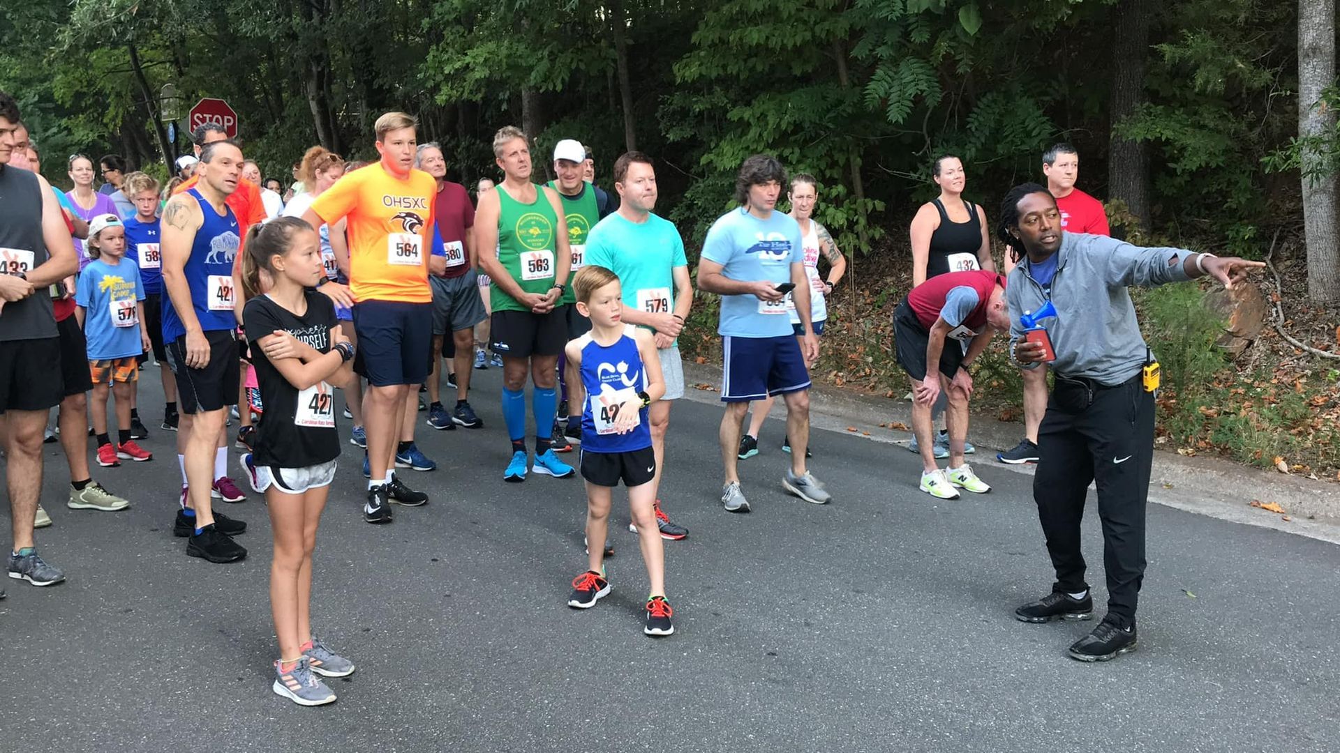 Runner in blue shirt leads a group stretching on a road beside trees.