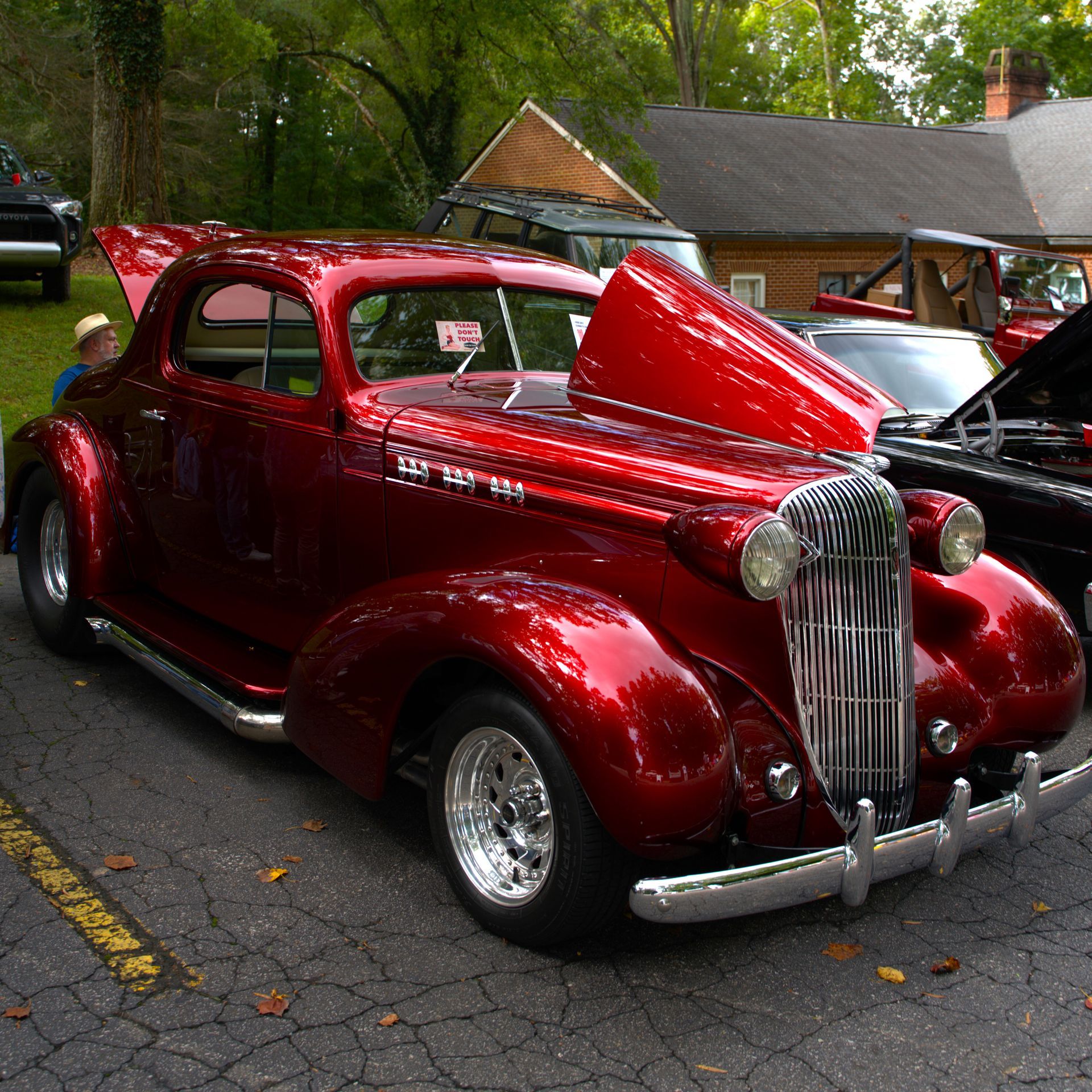 Shiny red vintage coupe at a car show, parked on wet pavement with its hood open.