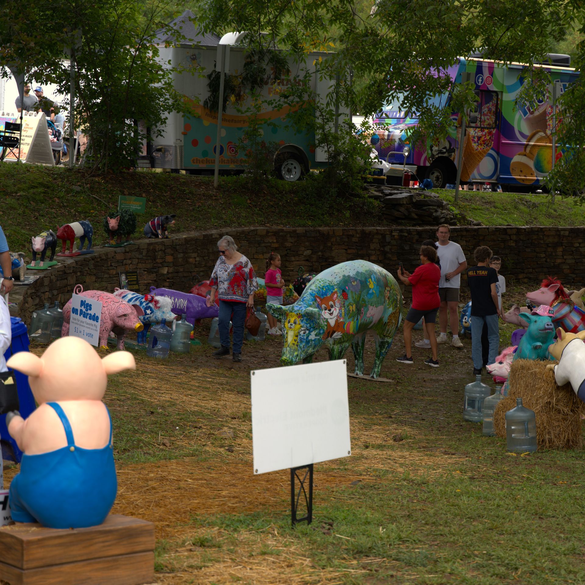 Outdoor fair scene with people gathered around colorful art displays and a blue pig statue on a grassy hillside