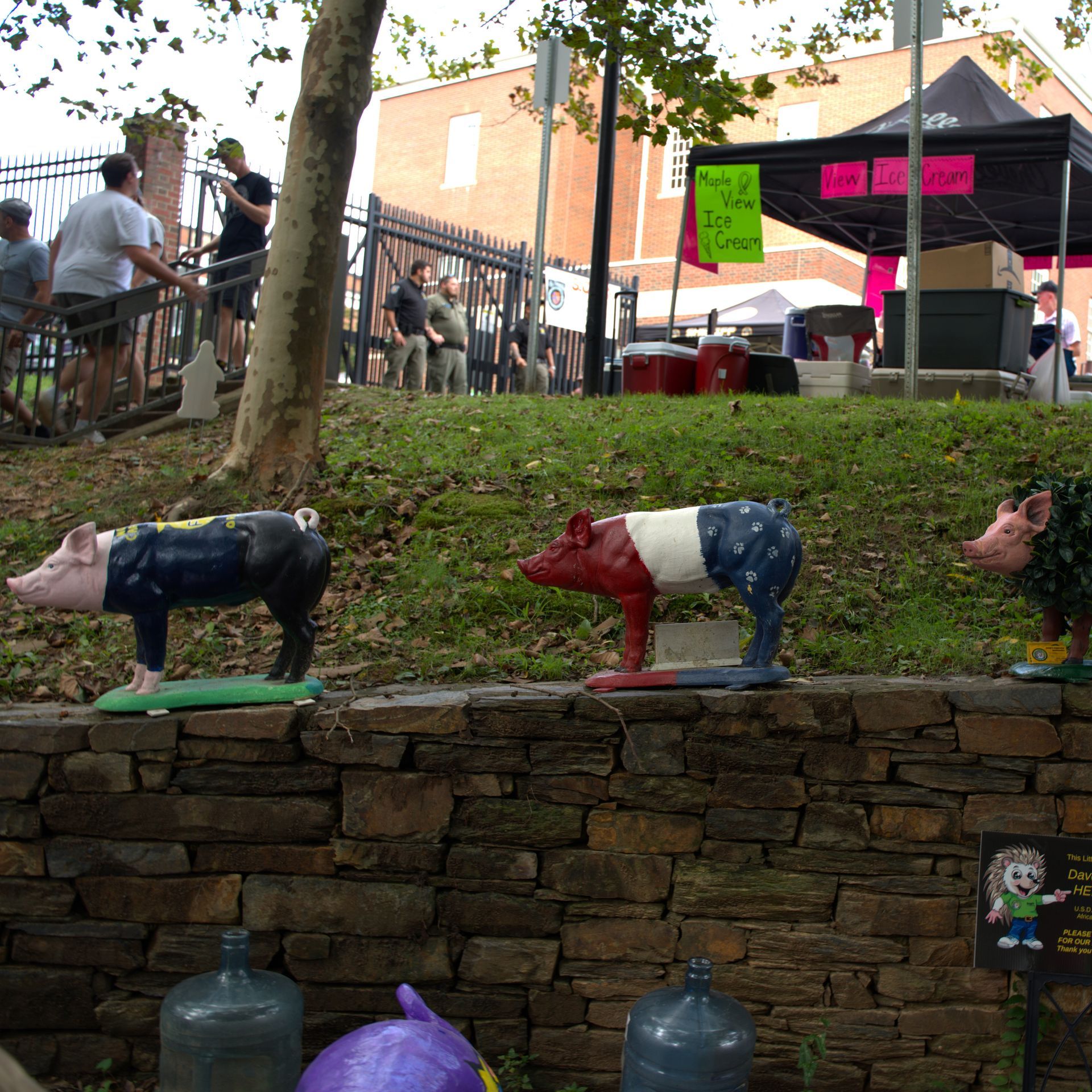 Three painted pig statues on a stone wall at an outdoor fair with tents and people in the background