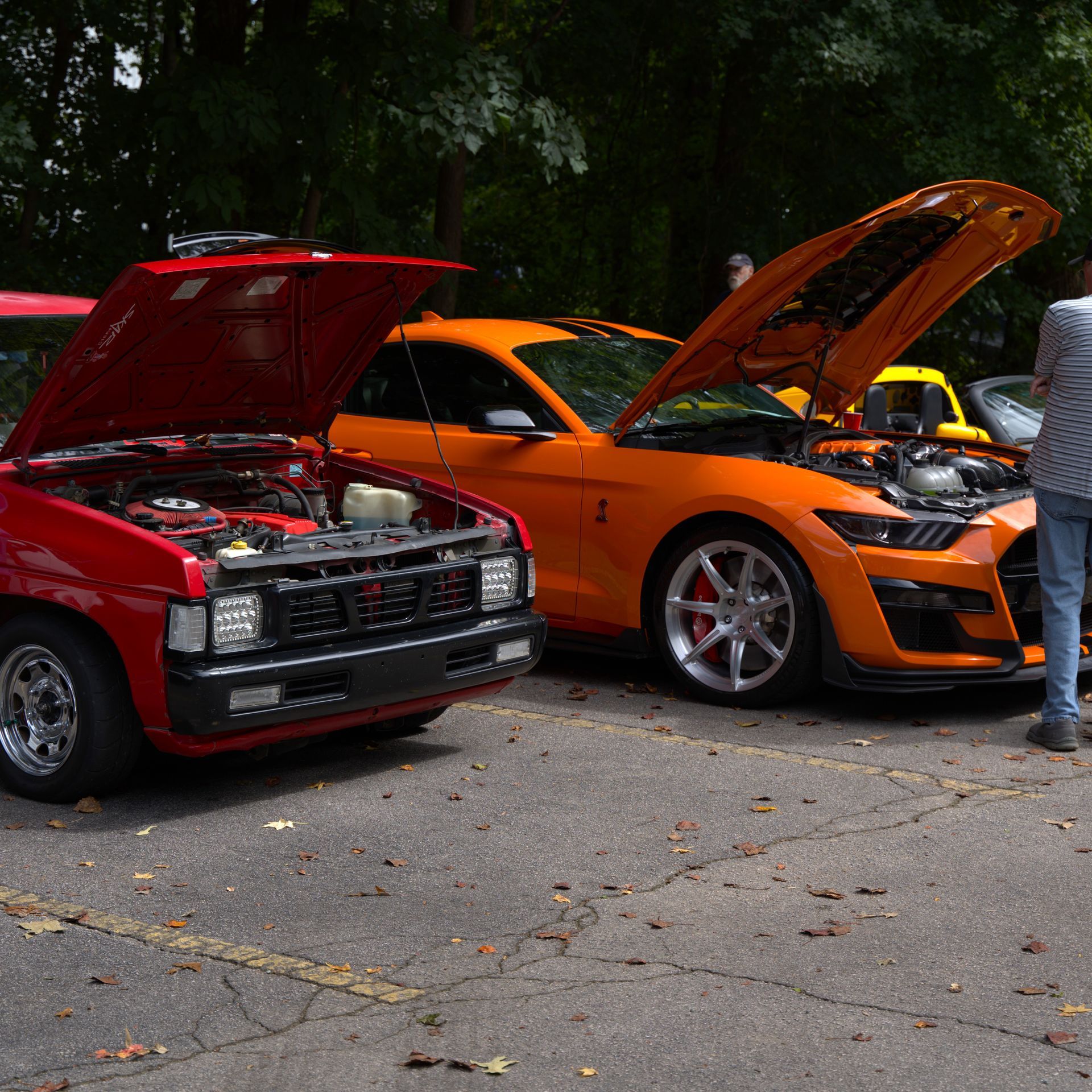 Two classic cars with open hoods at a car show, one red and one orange, with people nearby