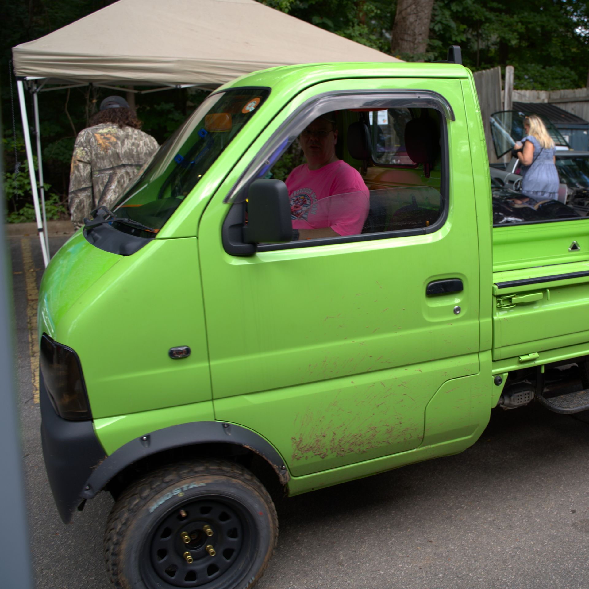 Bright green mini pickup truck at an outdoor market, with people and a tent in the background