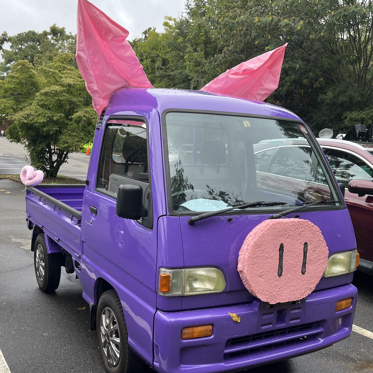 Purple mini truck decorated like a pig with pink ears and snout, parked outdoors on a rainy day