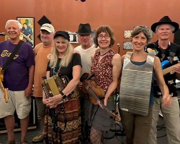 Six musicians stand indoors holding string instruments, smiling in front of a warm orange wall.
