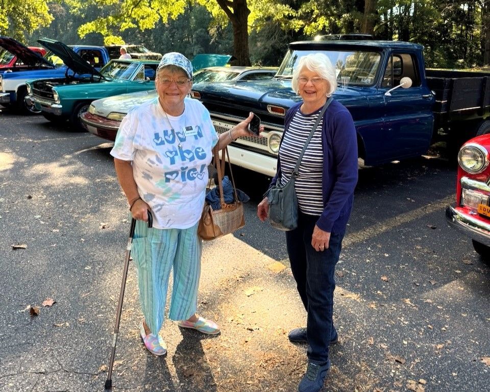 Two smiling people standing by classic cars on a sunny street, one holding a basket.