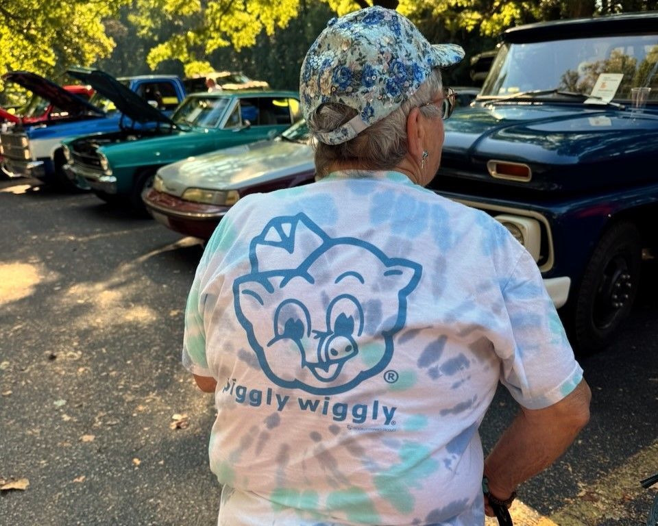 Person in a tie-dye Wiggy Wiggy shirt and camo cap stands by classic cars in a sunny parking lot.
