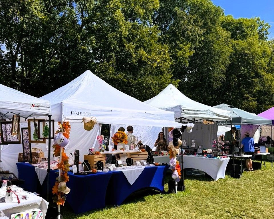 Outdoor craft fair with white tents, tables of handmade goods, and people browsing on a sunny grassy field