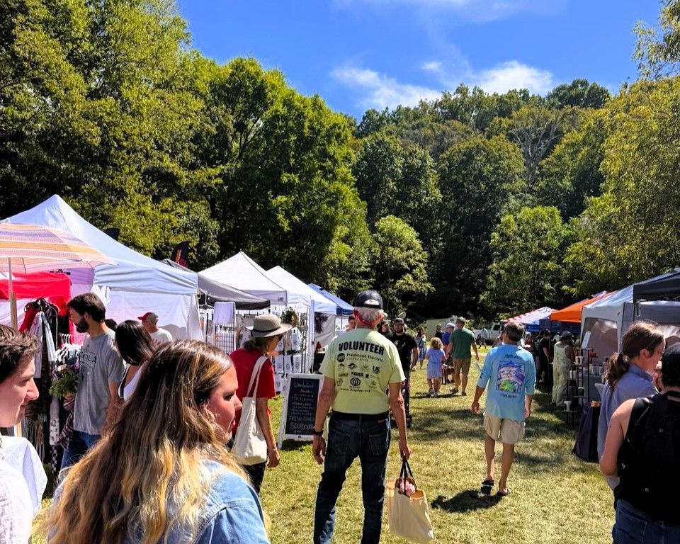 Outdoor market with white tents, people walking on a sunny grassy field, and trees in the background