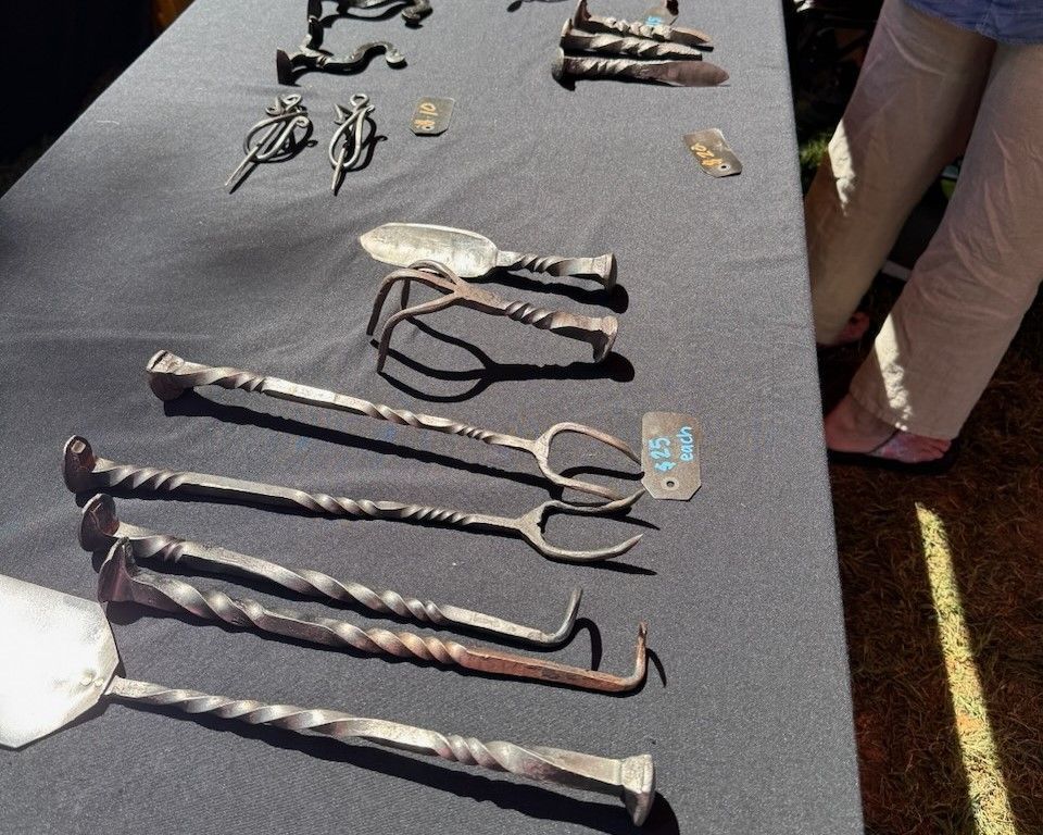 Assorted handmade metal tools and hooks laid out on a dark table at an outdoor market.