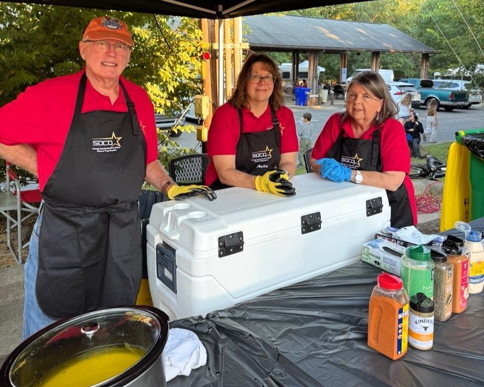 Three volunteers behind a table with coolers and drinks at an outdoor event.